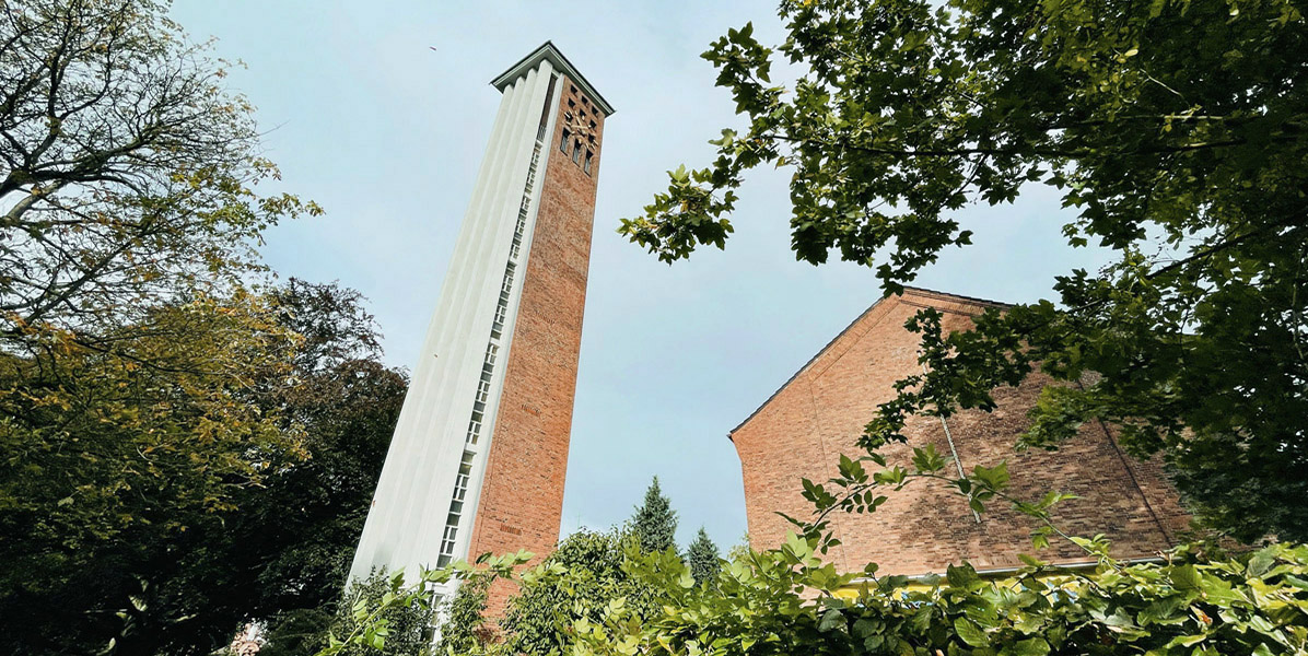 Ein hoher, schmaler Glockenturm aus Backstein mit vertikalen Fenstern steht neben einem Backsteingebäude, umgeben von grünen, belaubten Bäumen unter einem bewölkten Himmel.