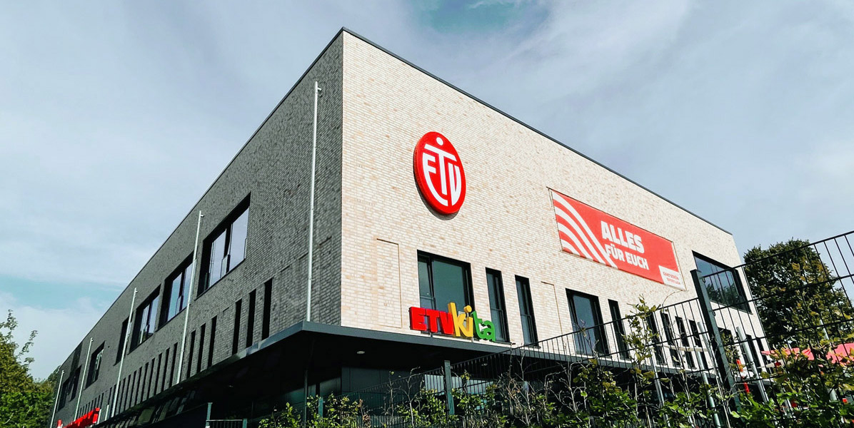 Ein modernes Gebäude mit hellen Backsteinwänden zeigt ein rotes FCI-Logo, ein rotes Banner mit der Aufschrift ALLES FÜR EUCH und ein buntes ETNika-Schild. Im Hintergrund sind Bäume und ein teilweise bewölkter Himmel zu sehen.