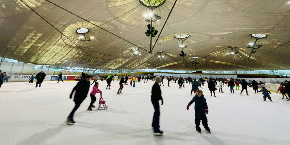 Eislaufende Menschen in einer großen, überdachten Eishalle mit einer hohen, zeltartigen Decke. Auf der Eisbahn herrscht reges Treiben mit Schlittschuhläufern verschiedenen Alters, einige mit Eislaufhilfen, bei warmer gelber Beleuchtung.