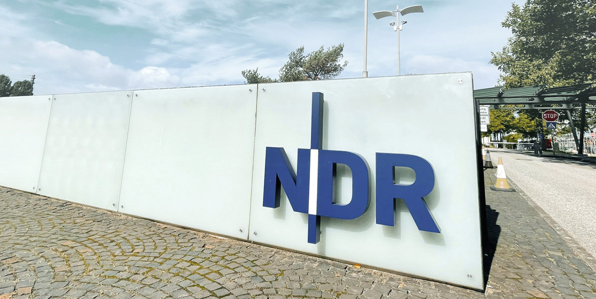 Ein großes Schild mit den blauen Buchstaben NDR steht vor einer Glaswand auf einem kopfsteingepflasterten Bürgersteig, im Hintergrund sind Bäume und Straßenschilder zu sehen, der Himmel ist teilweise bewölkt.