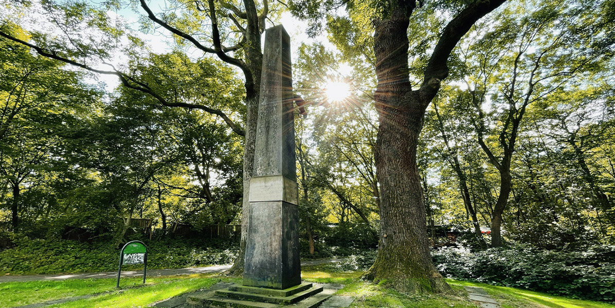 Ein hoher steinerner Obelisk steht in einem grünen, von Bäumen umgebenen Park. Das Sonnenlicht fällt durch die Äste und wirft einen hellen Schein und Schatten auf das Gras und das Denkmal.