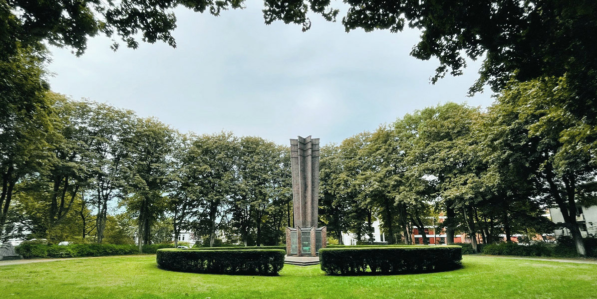 Ein hohes, schlankes Denkmal steht in der Mitte einer kreisförmig geschnittenen Hecke, umgeben von saftig grünem Gras und hohen Bäumen unter einem wolkenverhangenen Himmel, wobei die Gebäude teilweise durch die Bäume sichtbar sind.