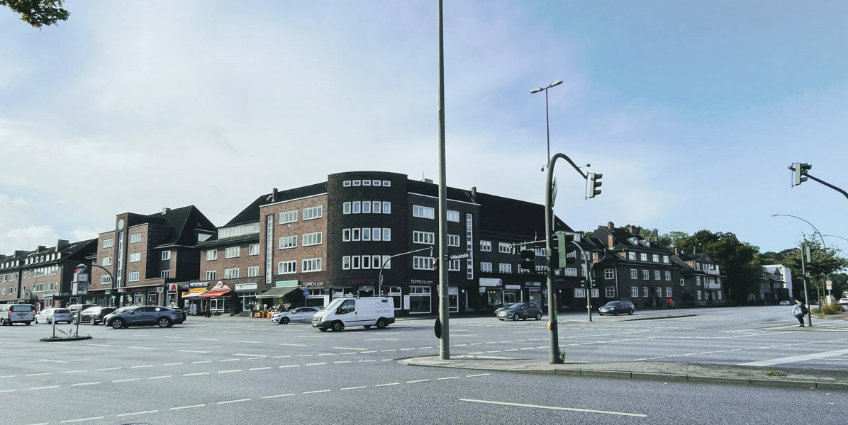 Eine breite, leere Kreuzung mit wenigen Fahrzeugen und einem großen, mehrstöckigen Backsteingebäude mit Geschäften und Restaurants im Erdgeschoss unter einem strahlend blauen Himmel. Ampeln und Fußgängerüberwege sind sichtbar.