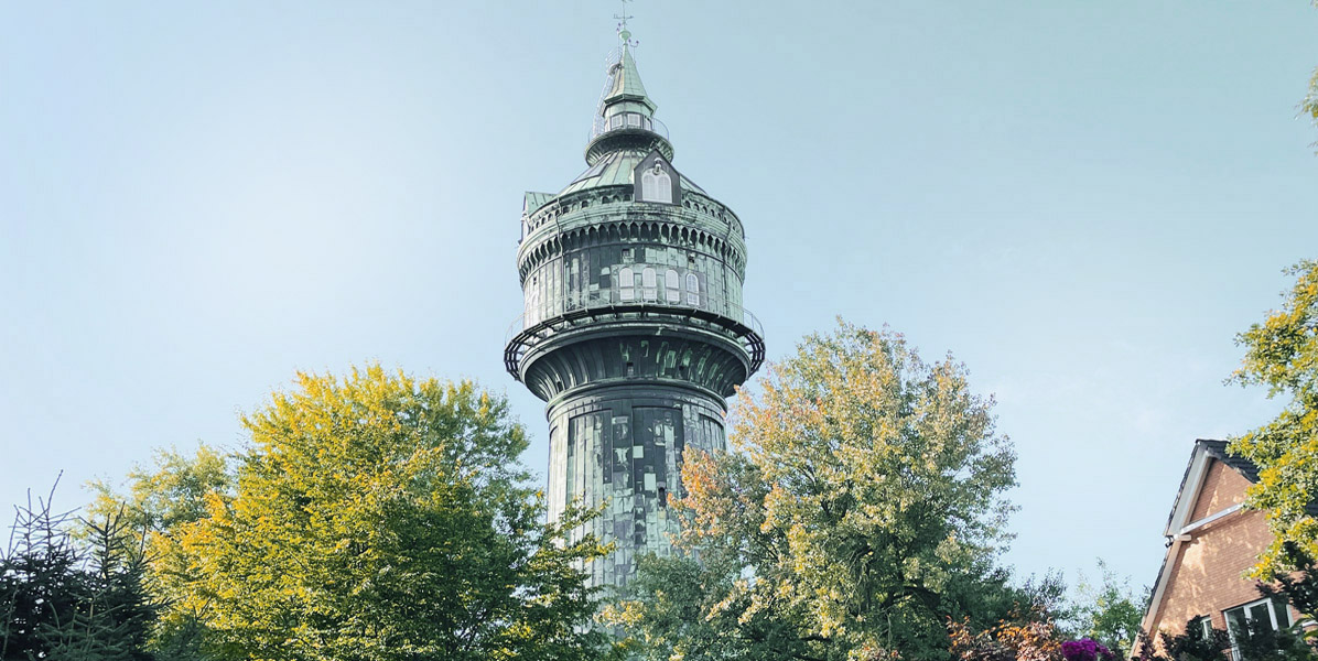 Ein hoher, historischer Wasserturm mit runder, verzierter Spitze erhebt sich über grünen und gelben Bäumen, wobei rechts ein Teil eines Gebäudes gegen den klaren Himmel zu sehen ist.