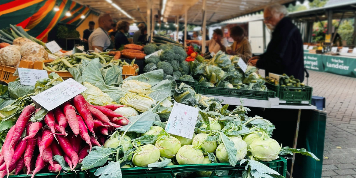 Frisches Gemüse, darunter rote Radieschen, Kohl, Brokkoli und Karotten, wird an einem Stand auf einem Bauernmarkt im Freien angeboten. Mehrere Personen kaufen ein und unterhalten sich im Hintergrund.