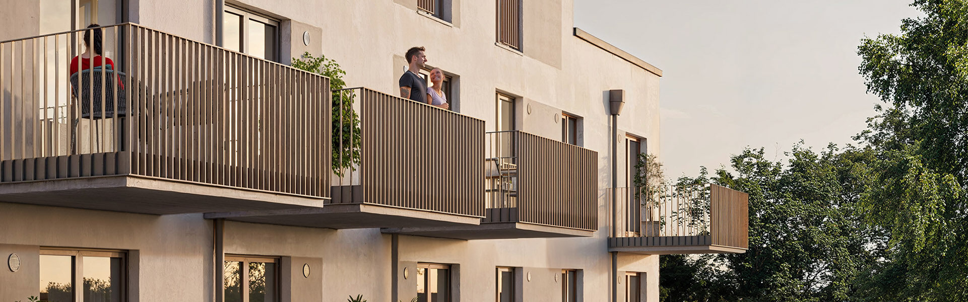 Rückansicht des modernen Gebäudes mit Wohnungen zum Kauf mit heller Fassade, großen Fenstern, gemeinschaftlichem Garten, Balkonen und Terrassen in der Abendsonne