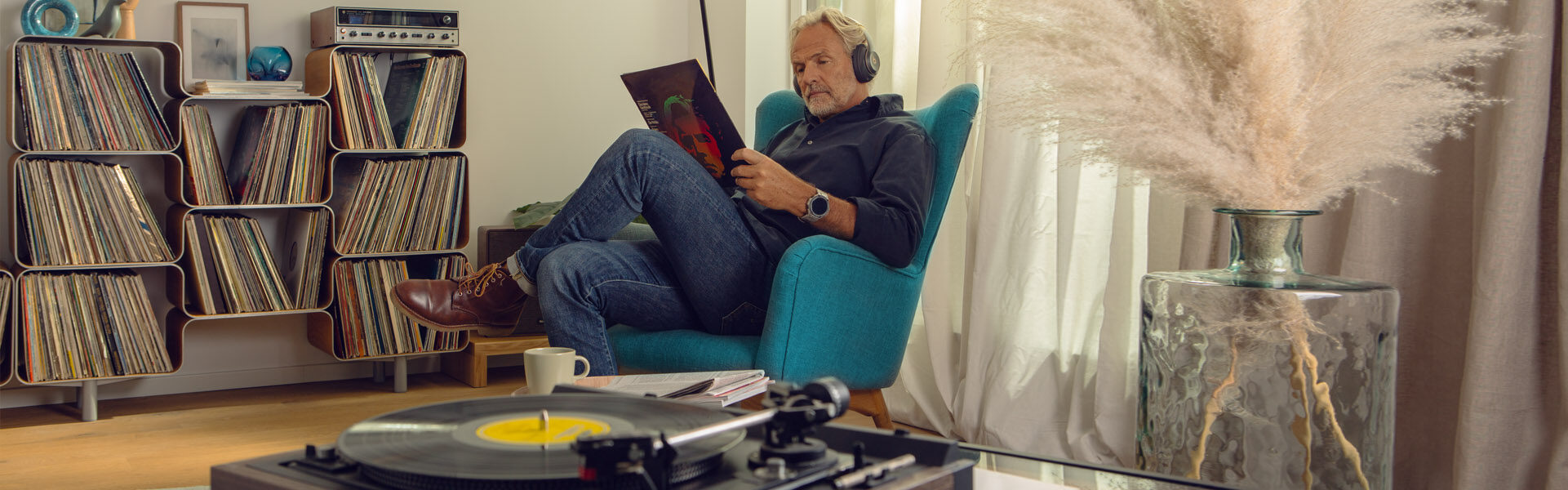 Ein Mann sitzt in einem blauen Stuhl, hört Musik über Kopfhörer und liest eine Schallplattenhülle. In der Nähe stehen Schallplatten und ein Plattenspieler. Durch das Fenster fällt Sonnenlicht und auf einem Tisch steht eine Vase mit Pampasgras.