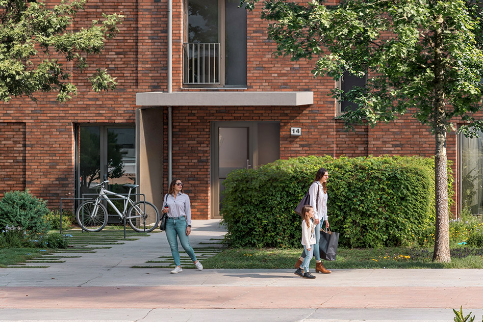 Bauprojekt mit Eogentumswohnungen zum Kauf von Behrendt mit überdachtem Eingang, Fahrradständer links, Hecken und Personen.