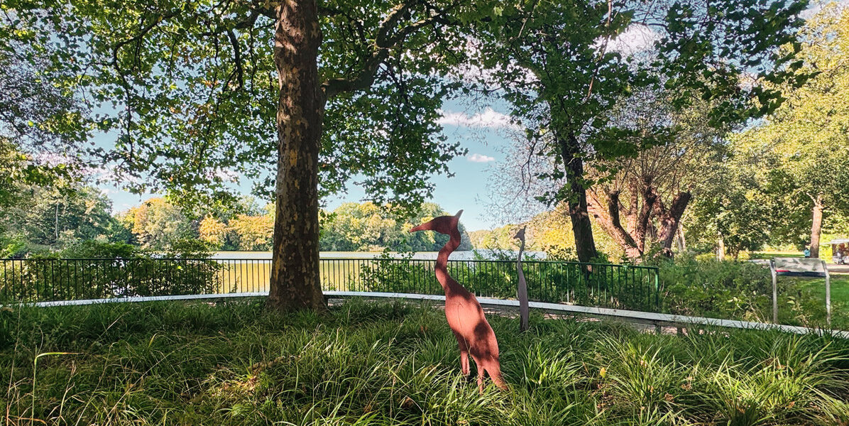 Eine rostfarbene Vogelskulptur steht inmitten von hohem, grünem Gras unter einem großen Baum, mit einem See und Bäumen im Hintergrund an einem sonnigen Tag in Hamburg.