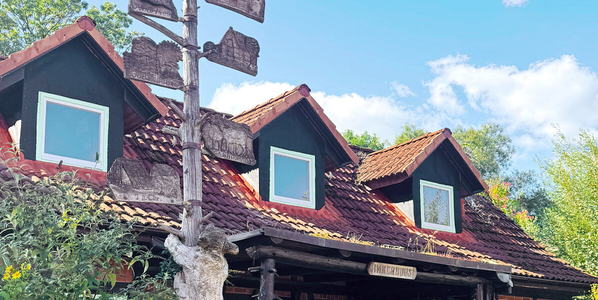 Ein rustikaler Holzbau mit rotem Ziegeldach und drei Dachgauben steht unter dem blauen Hamburger Himmel. Davor steht ein hoher Holzpfosten mit verwitterten, leeren Schildern - ein ziemlicher Kontrast zu jeder modernen Neubauwohnung in der Nähe.