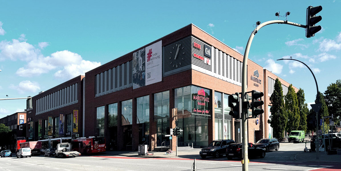 Eine moderne Neubauwohnung aus Backstein an einer Hamburger Stadtecke beherbergt ein Café und mehrere Geschäfte. Autos, Busse und Fußgänger sind zu sehen, mit Ampeln und Straßenschildern im Vordergrund. Der Himmel ist klar mit vereinzelten Wolken.