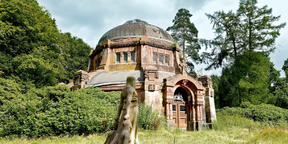 Ohlsdorfer Friedhof: Ein kuppelförmiges Steinmausoleum mit gewölbtem Eingang liegt inmitten von dichtem Grün und hohen Bäumen in Hamburg; im grasbewachsenen Vordergrund ist eine verwitterte Frauenstatue zu sehen, die den Kontrast zur modernen Neubauwohnung in der Nähe wiedergibt.
