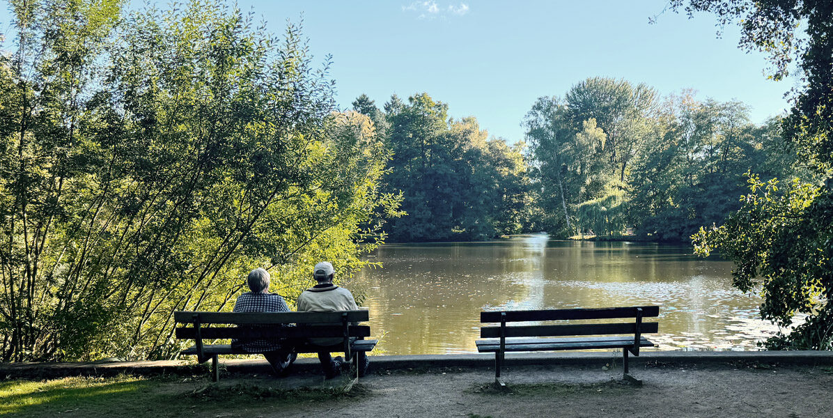 Zwei Menschen sitzen an einem sonnigen Tag auf einer Bank an einem friedlichen See in Hamburg, umgeben von Bäumen, mit einer leeren Bank in der Nähe und gedämpftem Sonnenlicht, das durch das Laub fällt - nur wenige Schritte von ihrer Neubauwohnung entfernt.