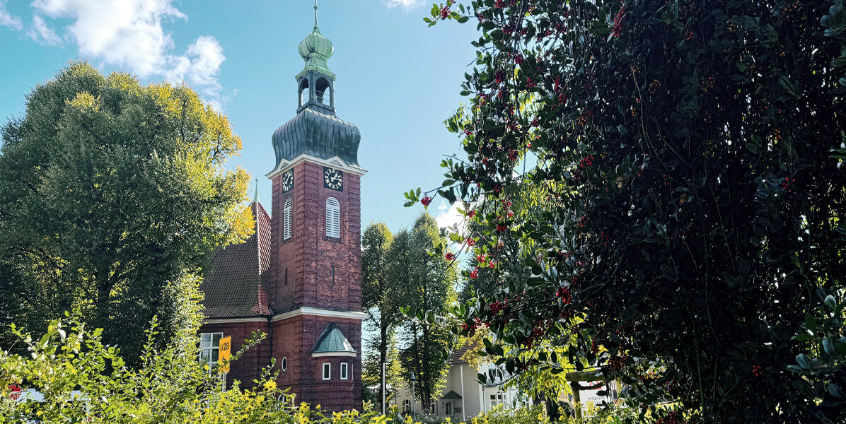 Eine rote Backsteinkirche mit Glockenturm und grünem Dach steht zwischen Bäumen und Büschen an einem sonnigen Tag in Hamburg, mit blauem Himmel und Wolken darüber. Zweige mit roten Beeren rahmen die rechte Seite ein, in der Nähe der Stelle, wo eine moderne Neubauwohnung stehen könnte.