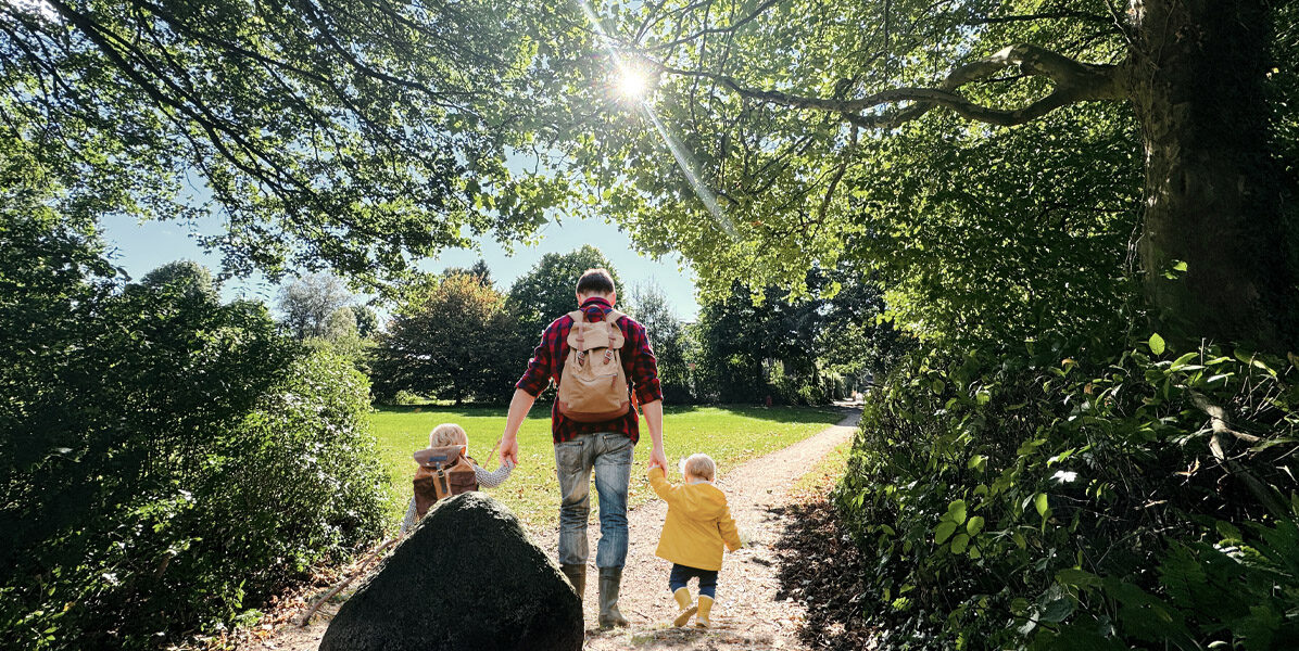Ein Erwachsener mit einem Rucksack geht auf einem sonnenbeschienenen Weg durch einen grünen Park in Hamburg und hält zwei kleine Kinder in bunten Jacken an der Hand. Bäume und Laub rahmen die Szene ein und weisen auf die nahe gelegenen Neubauwohnungen hin. Das Sonnenlicht fällt durch die Äste.