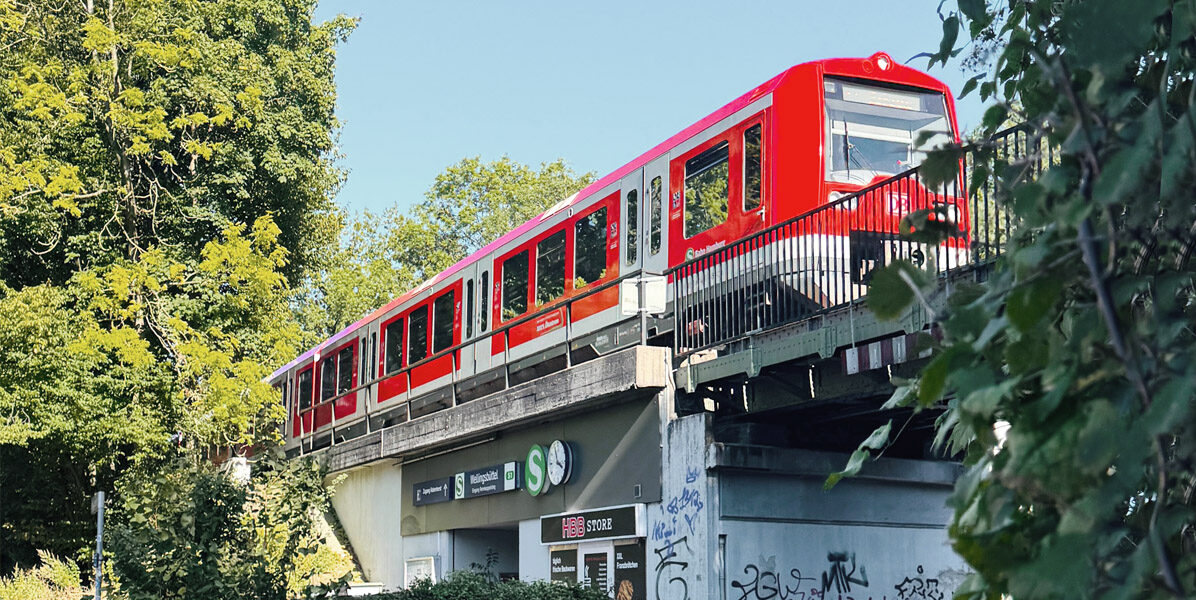 Ein leuchtend roter Zug fährt auf einer Brücke über einem kleinen Einkaufszentrum in Hamburg, umgeben von grünen Bäumen und blauem Himmel, mit Graffiti an der Wand unterhalb der Brücke.