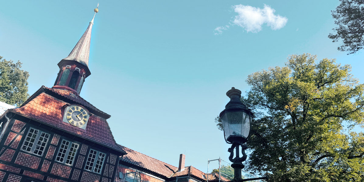 Ein historisches Backsteingebäude mit Uhrenturm und Kirchturm steht neben einer hohen Straßenlaterne und belaubten Bäumen in Hamburg, unter einem klaren blauen Himmel mit einer kleinen weißen Wolke.