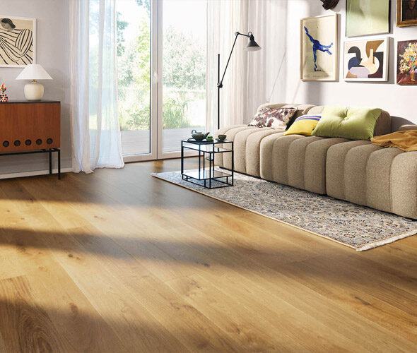 Ein helles Wohnzimmer in einer Neubauwohnung in Hamburg verfügt über Holzböden, ein beigefarbenes, geschwungenes Sofa mit bunten Kissen, einen gemusterten Teppich, ein modernes Sideboard, Wandkunst und große Glastüren, die natürliches Licht hereinlassen.