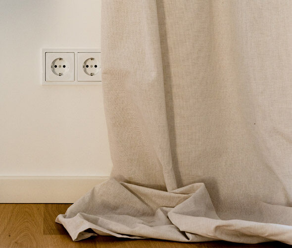 Ein beiger Vorhang verdeckt teilweise eine weiße Wand mit zwei Steckdosen im europäischen Stil in einer Hamburger Neubauwohnung. Der Vorhang fällt auf einen hellen Holzboden.