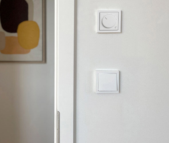 An einer weißen Wand in einer Hamburger Neubauwohnung befindet sich ein quadratischer Lichtschalter und darüber ein kreisförmiger Thermostat. Auf der linken Seite des Bildes ist ein Teil eines abstrakten Kunstwerks in gedeckten Farben zu sehen.