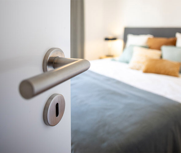 Eine teilweise geöffnete weiße Tür mit einem modernen silbernen Griff gibt den Blick frei auf ein gemütliches Schlafzimmer in einer Hamburger Neubauwohnung, in dem ein ordentlich gemachtes Bett mit einer grauen Decke und mehreren bunten Kissen steht.