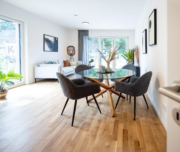 Helle, moderne Neubauwohnung in Hamburg mit hellen Holzböden, einem Glas-Esstisch mit schwarzen Stühlen, einem weißen Sofa, großen Fenstern, Topfpflanzen und minimalistischem Wanddekor.