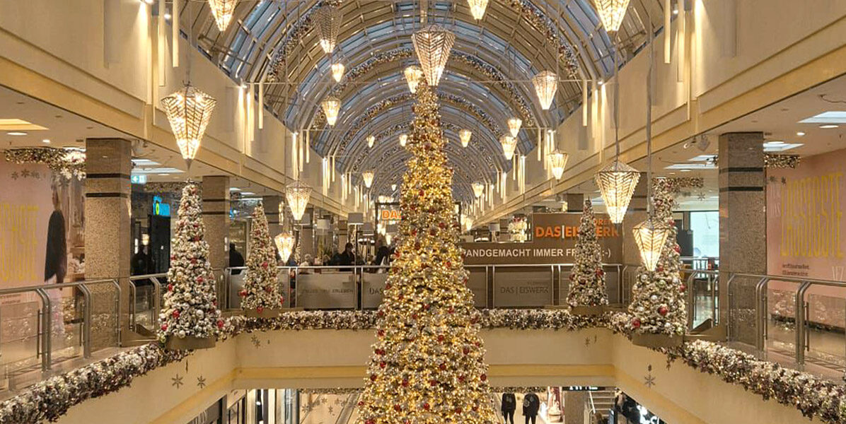 Alstertaler Einkaufszentrum (AEZ): Ein großes Einkaufszentrum in Hamburg ist weihnachtlich geschmückt. In der Mitte steht ein großer, geschmückter Weihnachtsbaum, umgeben von festlichen Lichtern und Girlanden. Auf beiden Ebenen des hell erleuchteten Einkaufszentrums flanieren die Menschen und genießen die Auslagen der Neubauwohnungen.