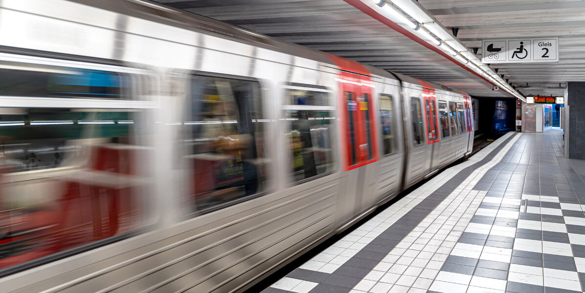 Ein silberner und roter U-Bahn-Zug rast durch einen unterirdischen Hamburger Bahnhof mit gefliestem Boden und Schildern über dem Kopf, darunter Symbole für Barrierefreiheit und Bahnsteig Nummer 2 - perfekt, um Ihre Neubauwohnung in der Stadt zu erreichen.